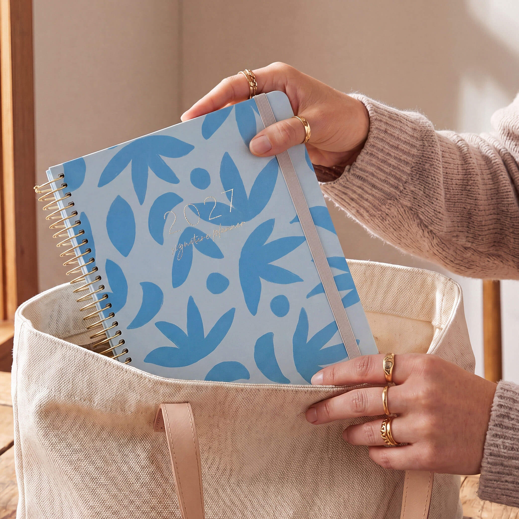 2027 Signature Planner in Sunburst Sky being placed into a canvas tote bag, gold coil and grey elastic closure.