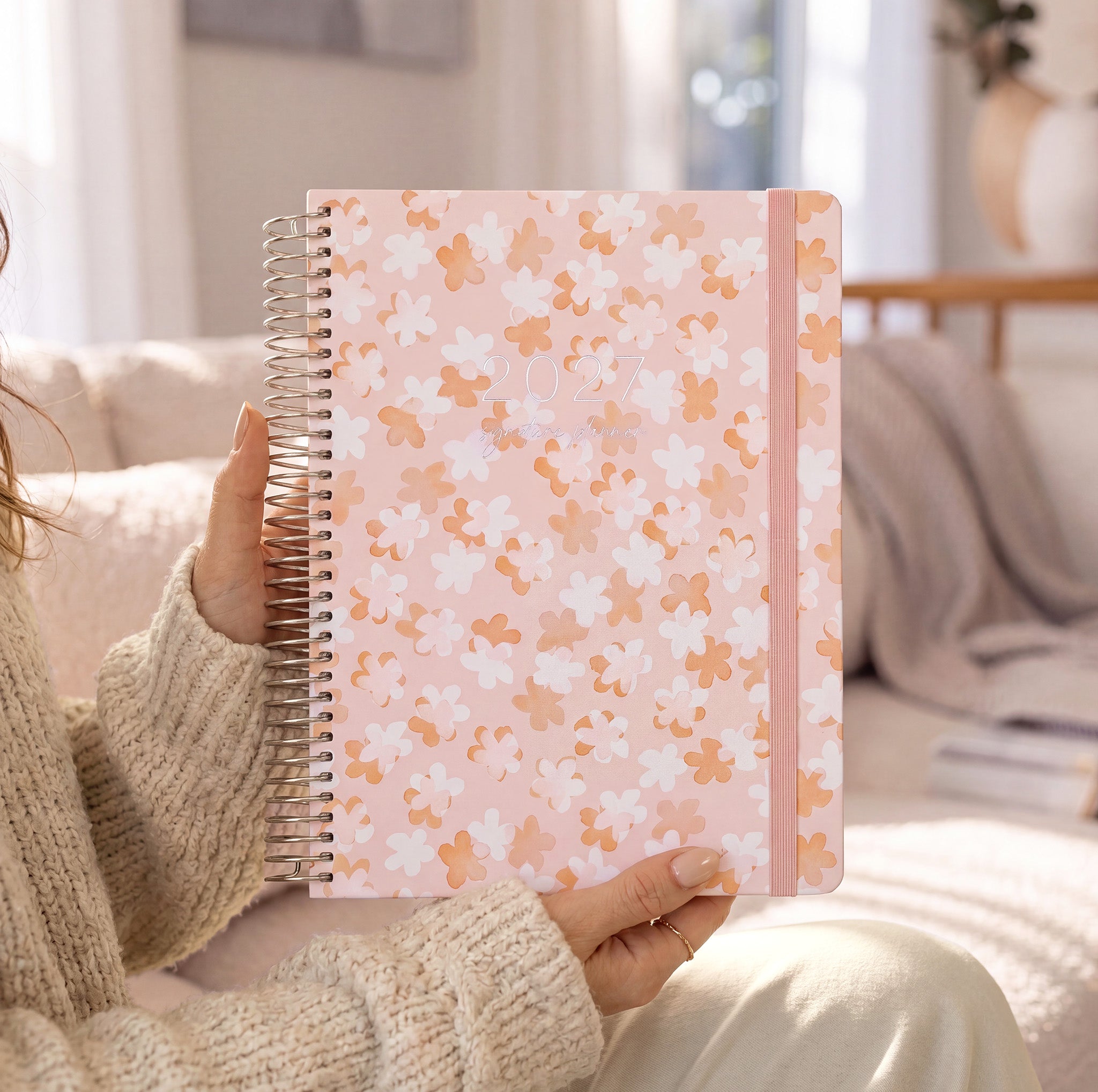 2027 Signature Planner in So Sweet held by a woman in a cream knit sweater, silver coil and pink elastic closure.