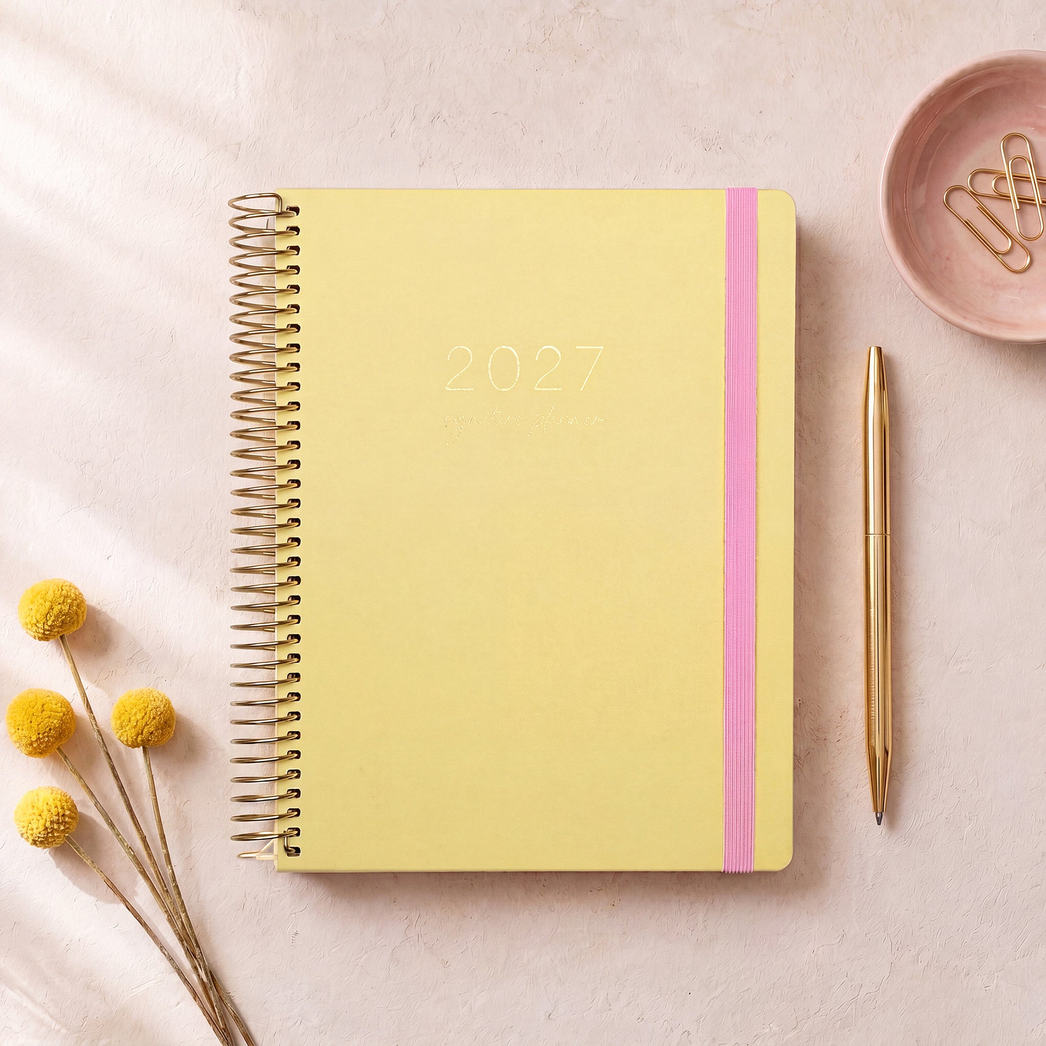 2027 Signature Planner in Butter styled with yellow dried florals and a gold pen, gold coil and pink elastic closure.