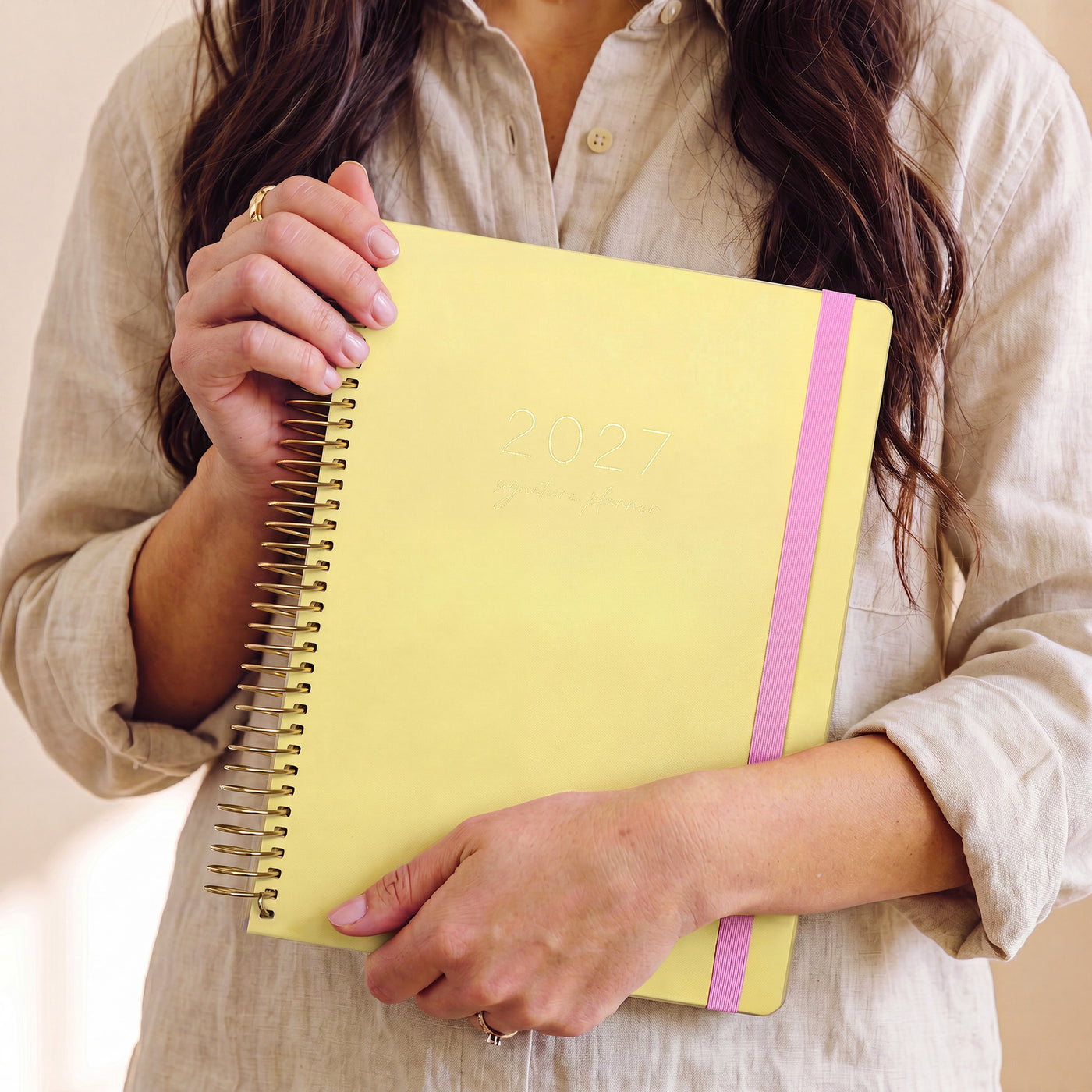2027 Signature Planner in Butter held by a woman in a linen shirt, gold coil and pink elastic closure.